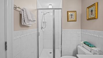 Un baño con ducha de vidrio, azulejos blancos, inodoro y cuadros en la pared.