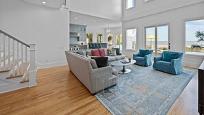 A spacious living room with a grey sectional sofa, two blue armchairs, a coffee table, and a patterned rug. A staircase with white railings is visible on the left. - Luxury Oceanfront- Private Pool, Hot Tub, & Elevator! Can't beat location! (Isle Of Palms)