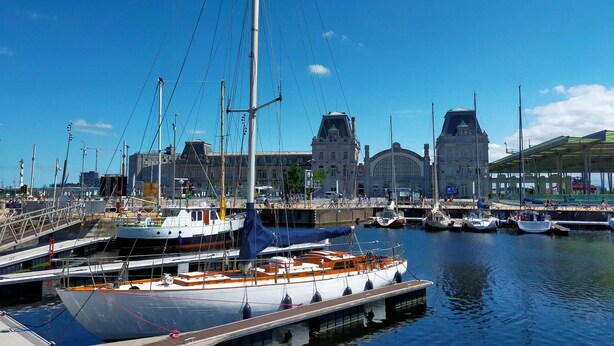 Un port de plaisance où sont amarrés plusieurs bateaux, un grand bâtiment avec un clocher en arrière-plan et un ciel bleu dégagé.