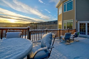 Un deck cubierto de nieve con mobiliario exterior y vista a un paisaje boscoso.