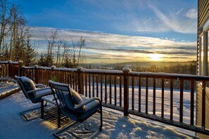 A snow-covered deck with two lounge chairs and a wooden railing overlooking a snowy landscape with a sunset.