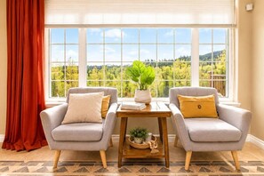 A room with two grey armchairs, a wooden table with a plant, and a view of a forest through large windows.
