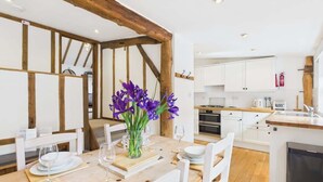 A dining area with a wooden table set for two, white chairs, and a vase of purple flowers. - The Cote, Little Upton Farm (Ashey)