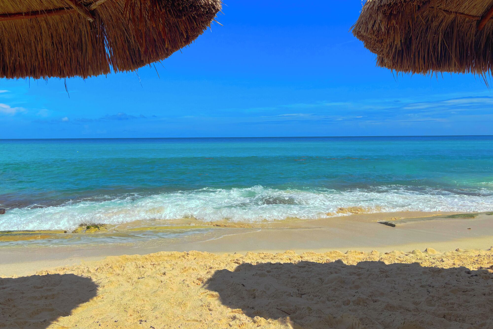 A beach with clear blue water, sandy shore, and thatched umbrellas.