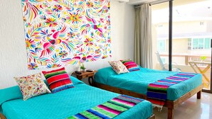 A room with two beds, each with a colorful blanket and pillows, a small table with a plant, and a large window with a view of buildings. - Renovated Studio in Cancun Beach (Cancún)