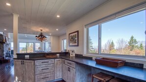 A spacious kitchen with a large island, wooden cabinets, and a view of trees through large windows. - Saddleback - Rock Pond Condo 24 - right above base lodge (Rangeley)