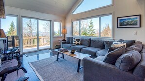 A spacious living room with a gray sofa, a coffee table, and a large window offering a view of trees and mountains. - Saddleback - Rock Pond Condo 24 - right above base lodge (Rangeley)