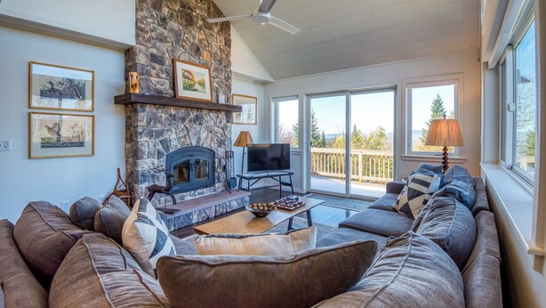 A cozy living room with a stone fireplace, comfortable seating, and large windows offering a view of the outdoors. - Saddleback - Rock Pond Condo 24 - right above base lodge (Rangeley)
