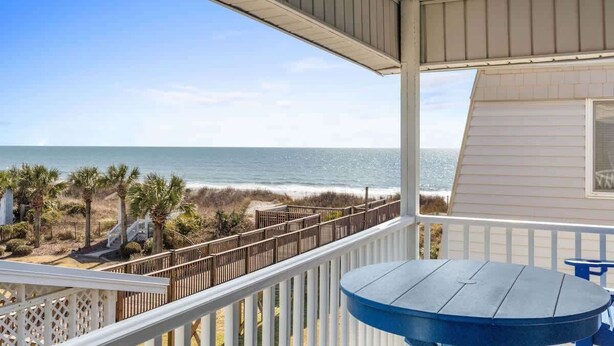 A balcony with a view of the ocean, a wooden table, and a white railing. - I'll Beach There For You by Brunswick VR: OIB Oceanfront w/ Pool & Water Views (Ocean Isle Beach)