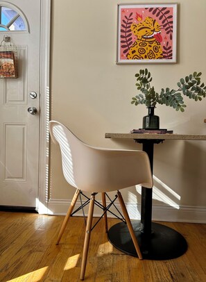 Une pièce avec une chaise blanche, une petite table sur laquelle se trouve une plante, et une œuvre d’art encadrée accrochée au mur.