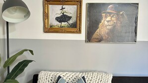 Een woonkamer met een donkere bank, een witte gebreide plaid en gestreepte kussens. Er hangen lijstwerken aan de muur, staat een vloerlamp en een pot met een plant.