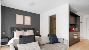 A modern bedroom with a bed, bedside tables, a grey sofa, and a kitchen area visible through an open door.
