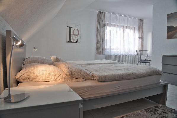 A bedroom with a bed, bedside lamps, a chair, a radiator, and a window with curtains.