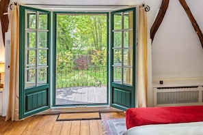 A room with a large open window, a wooden deck, and a bed with a red cover.