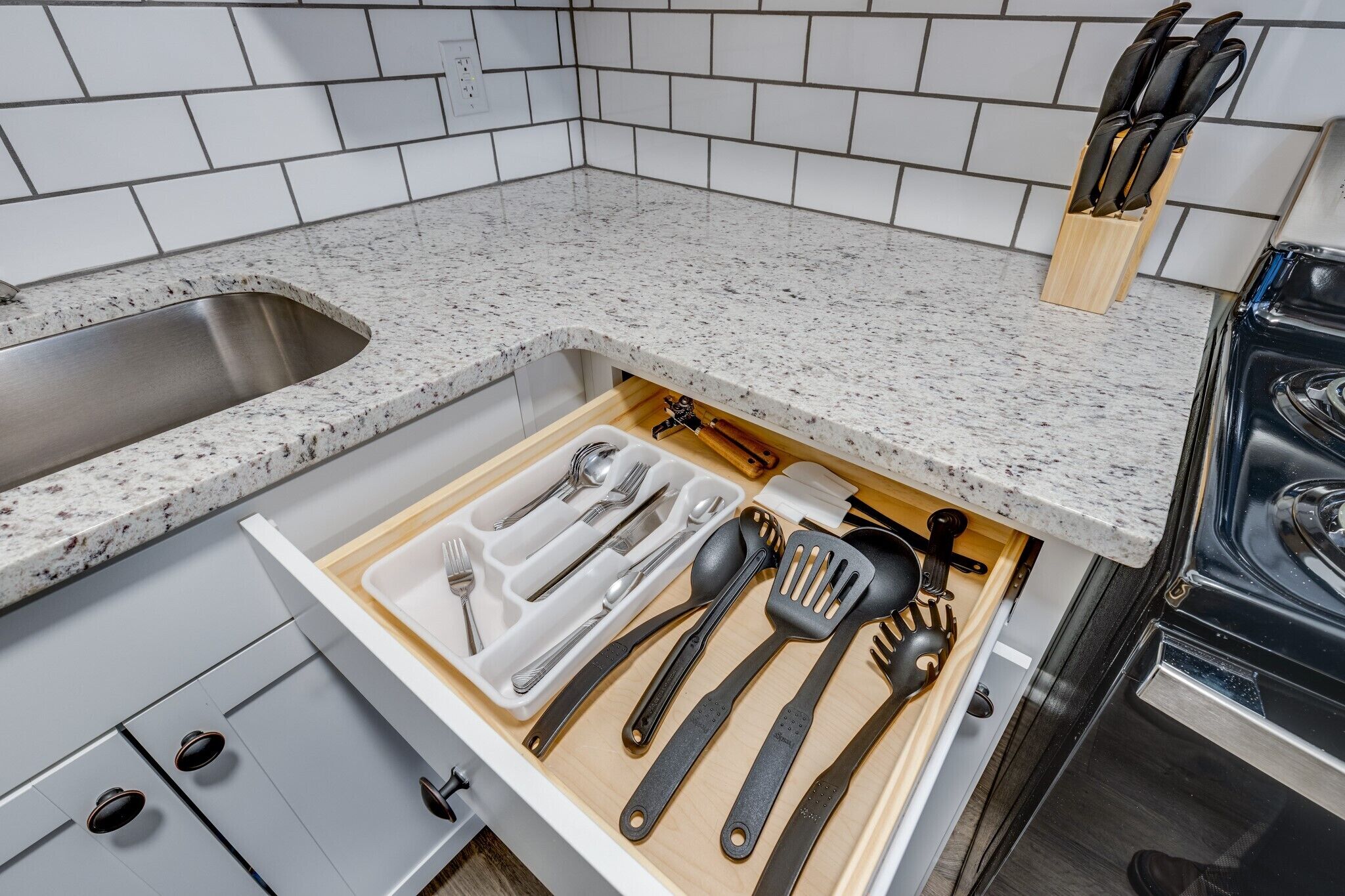 A kitchen with a granite countertop, a stainless steel sink, and a drawer organizer with utensils.