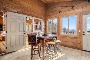Interior de una cabaña de madera con comedor, ventanas amplias y chimenea de piedra.