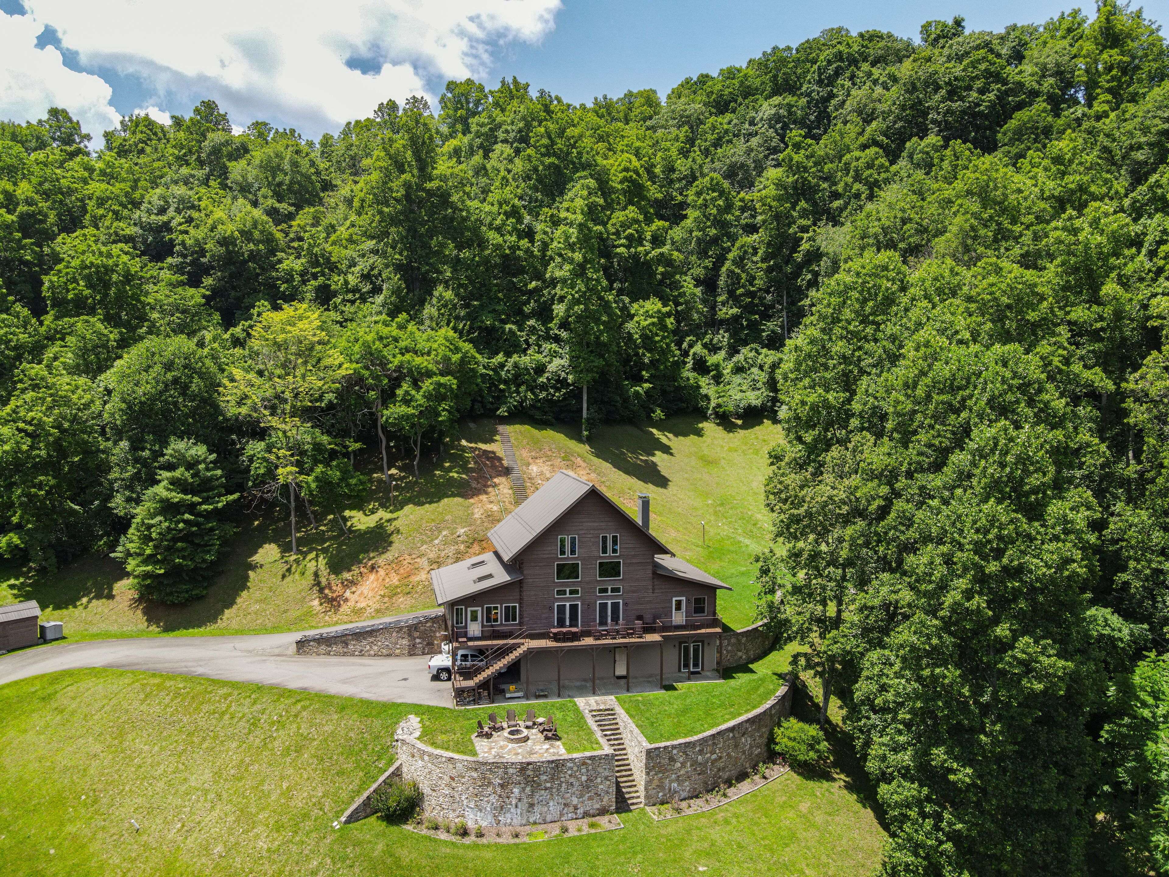 A large, two-story house with a stone foundation and a wrap-around porch, surrounded by dense forest.