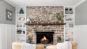Un salon chaleureux avec une cheminée en pierre, deux fauteuils beiges et une étagère blanche au-dessus de la cheminée, agrémentée d’objets décoratifs.