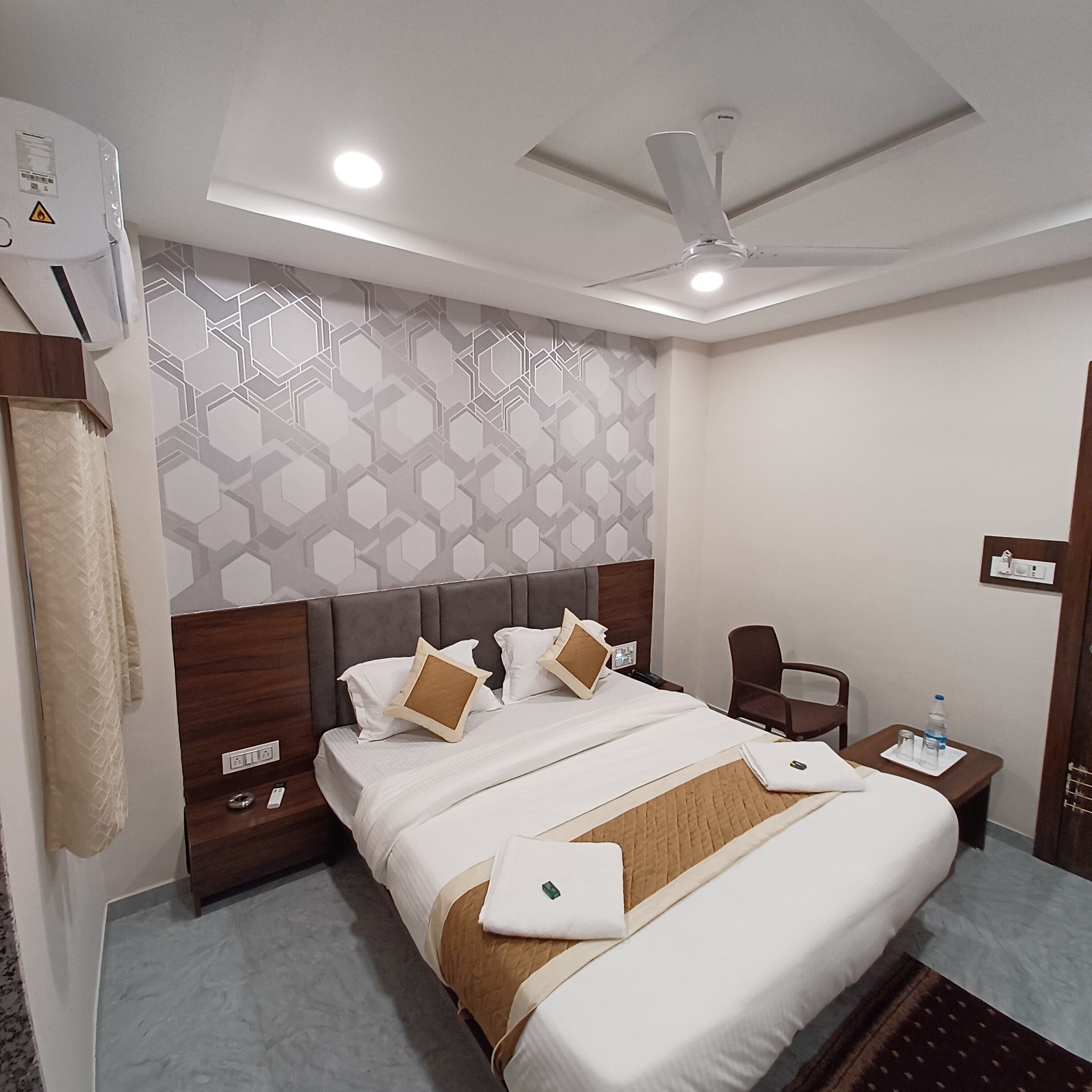 A hotel room with a large bed, a ceiling fan, a nightstand, a chair, and a wall with a geometric pattern.