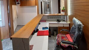 Zona de recepción de hotel con un mostrador de madera, un área de descanso con una silla, un ventilador de techo y un pasillo que conduce a otras habitaciones.