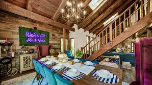 Un intérieur rustique en bois, agrémenté d’un lustre, d’un néon, d’un coin repas avec une table dressée pour quatre personnes et d’un escalier menant à un autre niveau.