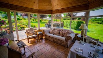 A conservatory with a wooden ceiling, a sofa, a dining table set, and a view of a garden.