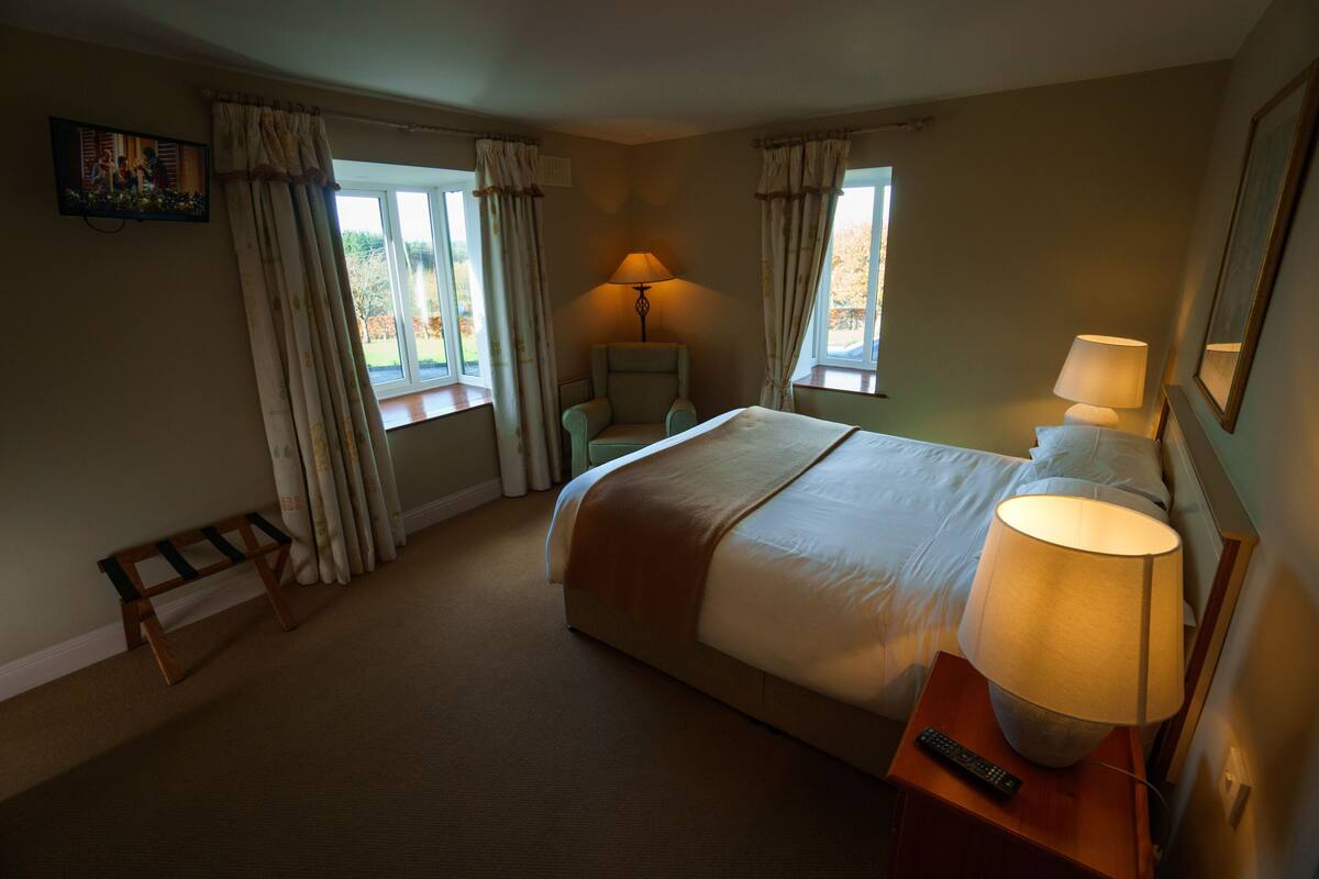 Quarto de hotel com cama, abajures, uma cadeira, uma mesinha e janela com cortinas.