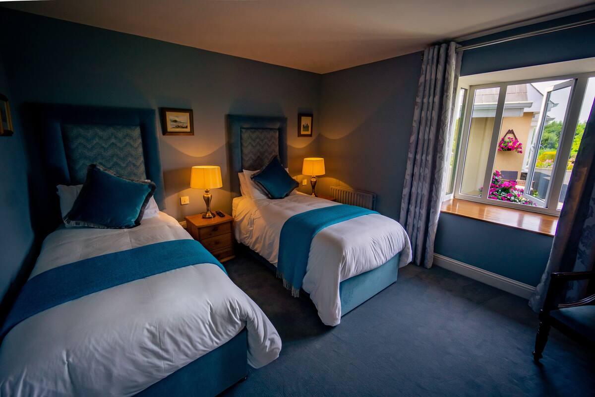 Quarto de hotel com duas camas, mesas de cabeceira com abajures, uma janela com vista para o exterior e uma cadeira.