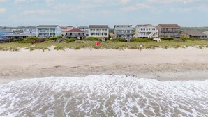 A beachfront property with multiple-story houses, a sandy beach, and a visible lifeguard flag. - Hobbs Realty - Oceanfront Home w/Expansive Decks and Second Living Area w/Futon (Holden Beach)