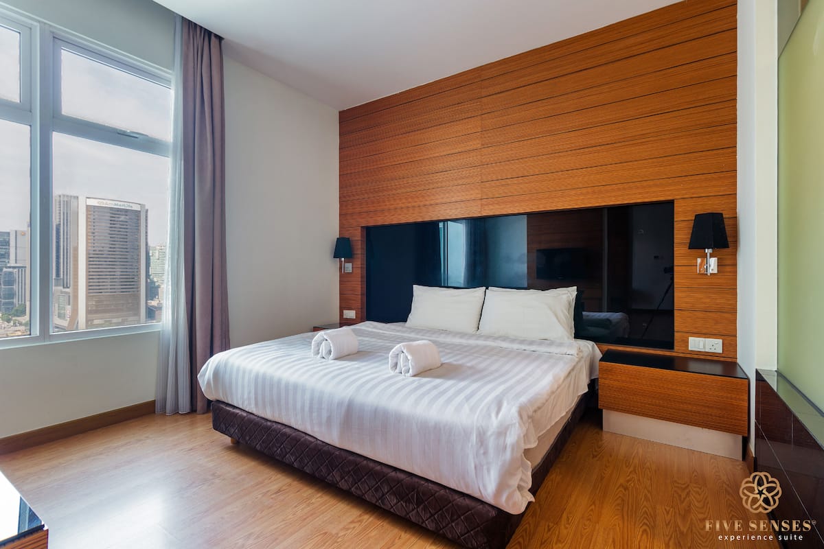 Ein modernes Hotelzimmer mit großem Bett, Holz-Kopfstütze, Blick auf die Stadt und einem Nachttisch mit Lampe.