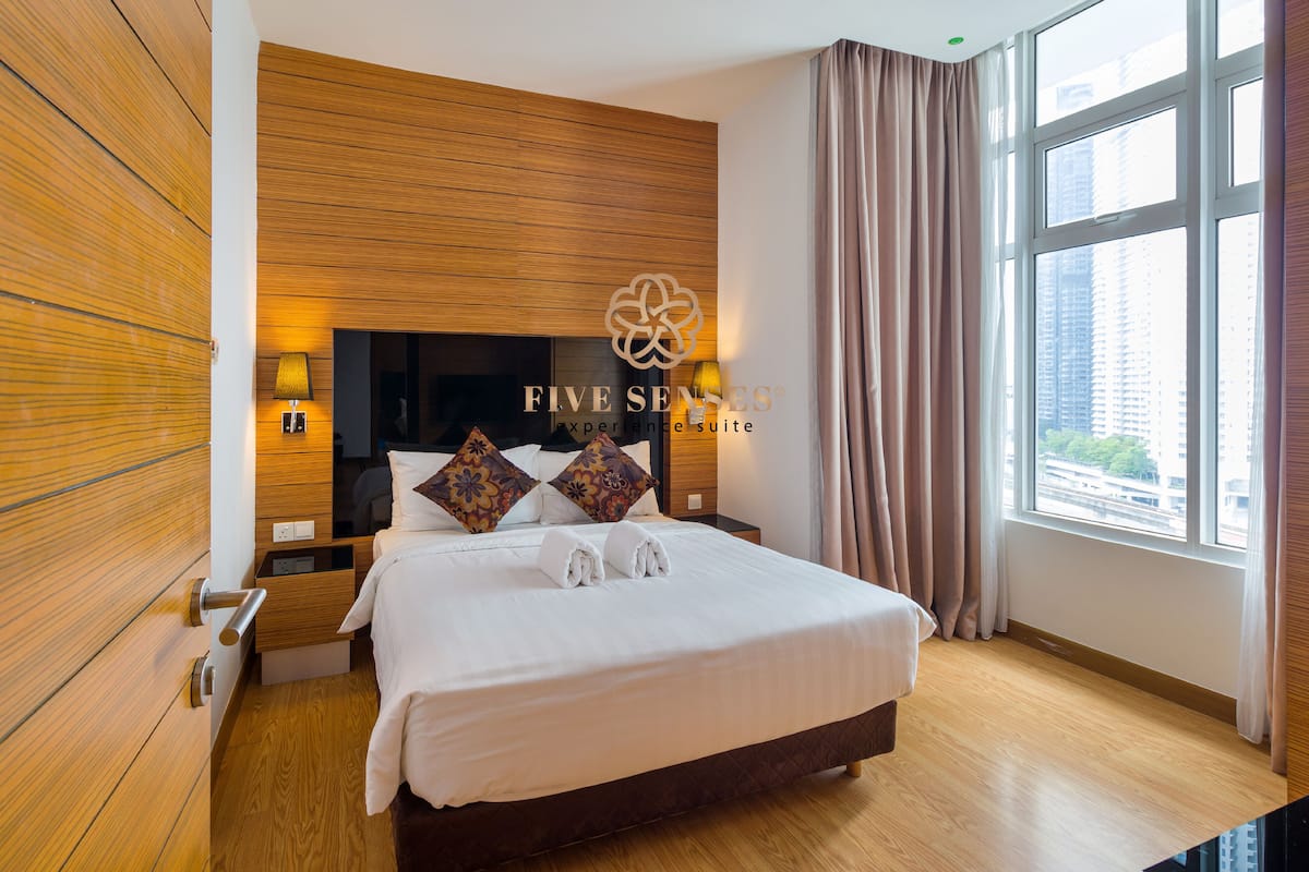 Ein modernes Hotelzimmer mit einem großen Bett, Holzvertäfelung, einem Nachttisch mit Lampe und Blick auf die Stadt durch ein großes Fenster.