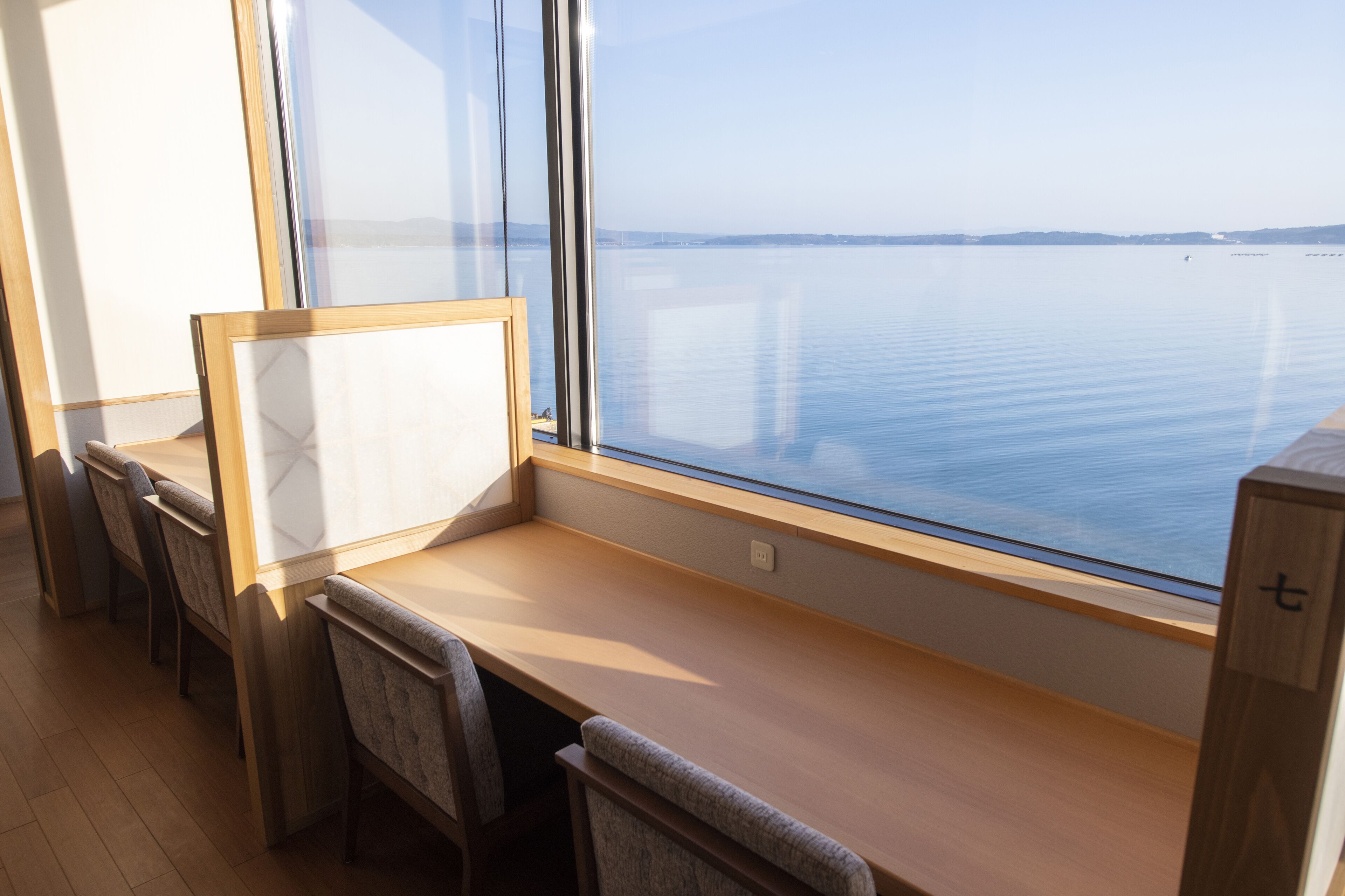 A room with a large window overlooking a calm body of water, a wooden desk, and several chairs.
