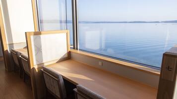 A room with a large window overlooking a calm body of water, a wooden desk, and several chairs.