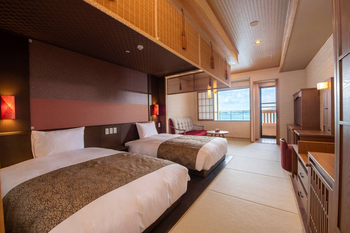 Habitación de hotel con dos camas, un rincón de descanso con una silla y mesa, y un balcón con vistas.