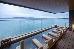 A modern infinity pool with wooden steps and railings, overlooking a calm body of water and distant land.