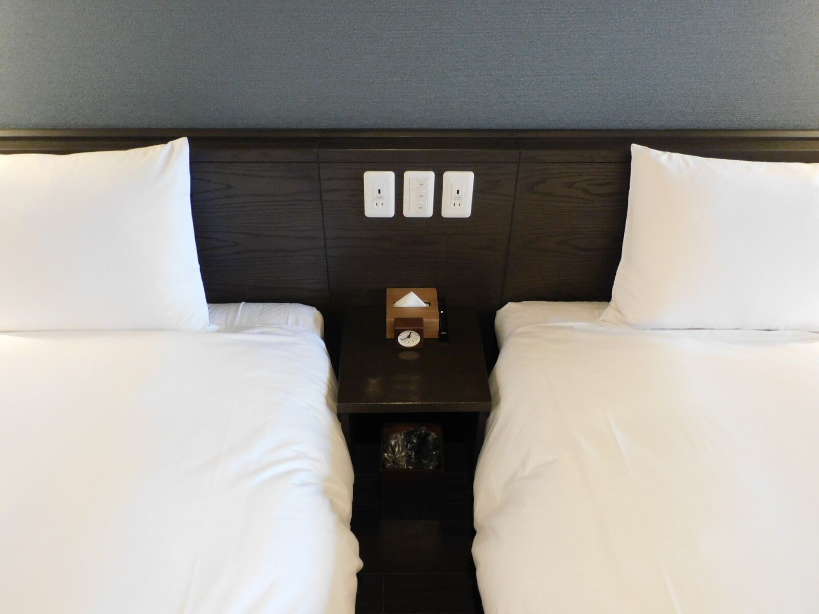A hotel room with two beds, a nightstand with a clock, and three electrical outlets on the wall.