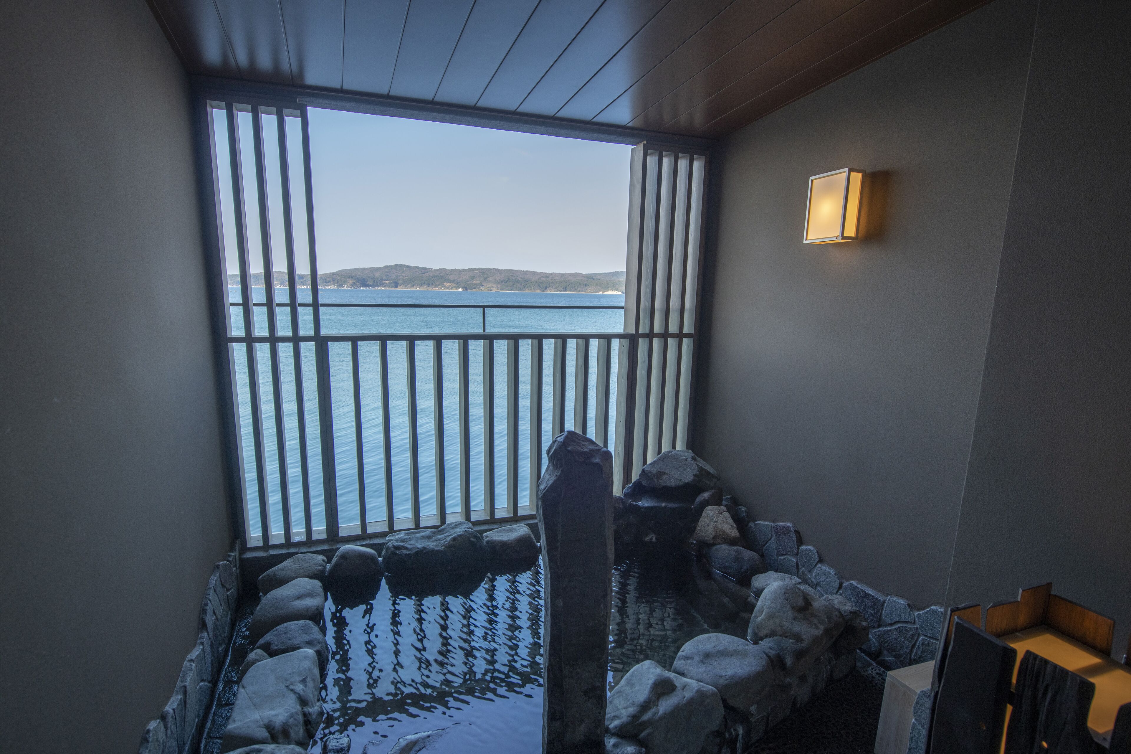 A room with a view of the sea, a stone wall, and a wooden ceiling.