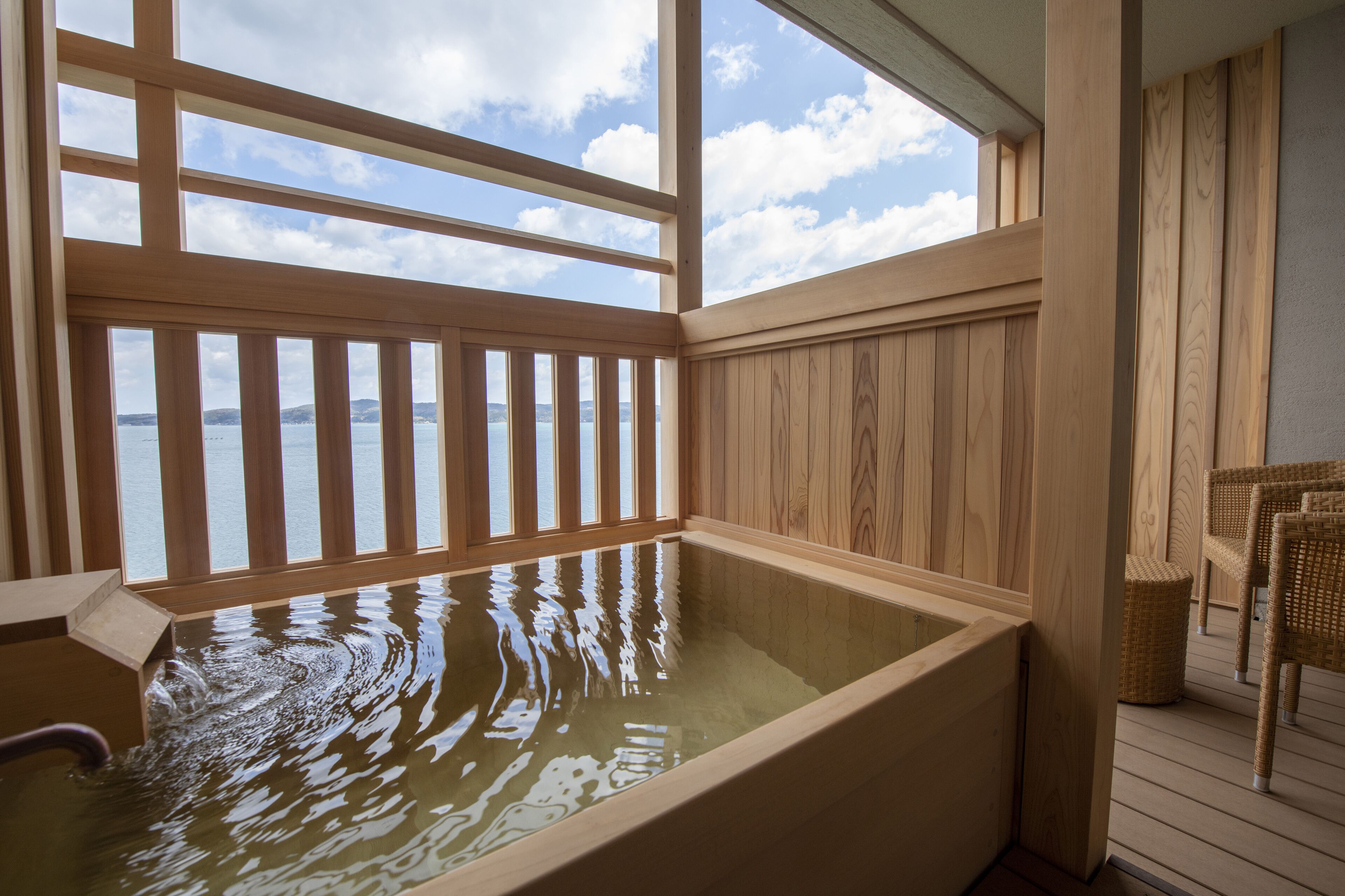 Una bañera de madera con vistas al mar, rodeada de paredes de madera y una silla de mimbre.