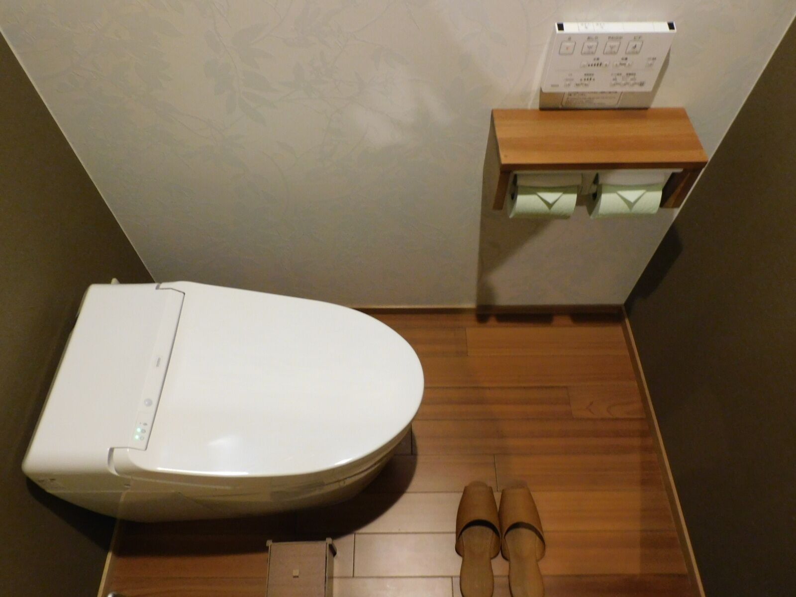 A small, compact bathroom with a white toilet, a wooden floor, and a small shelf with a box on top.