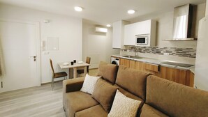 Apartament modern amb saló que disposa d'un sofà marró i una taula de dinar amb cadires. La cuina inclou microones, rentapesos i armaris de fusta.