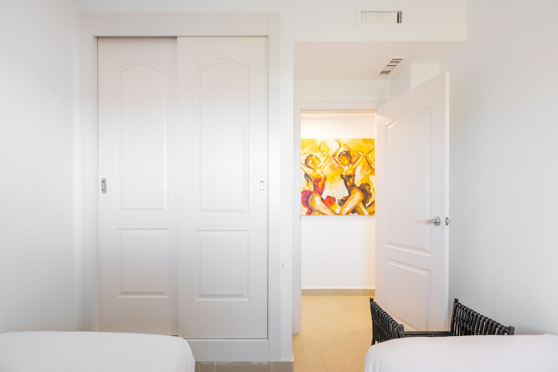 A hotel room with two beds, a white door, and a hallway leading to a bathroom with a colorful abstract painting.