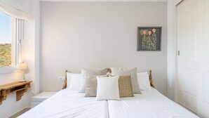 En pænt redt seng med hvide sengetæpper og mønstrede puder, et natbord med en lampe og et indrammet kunstværk på væggen.