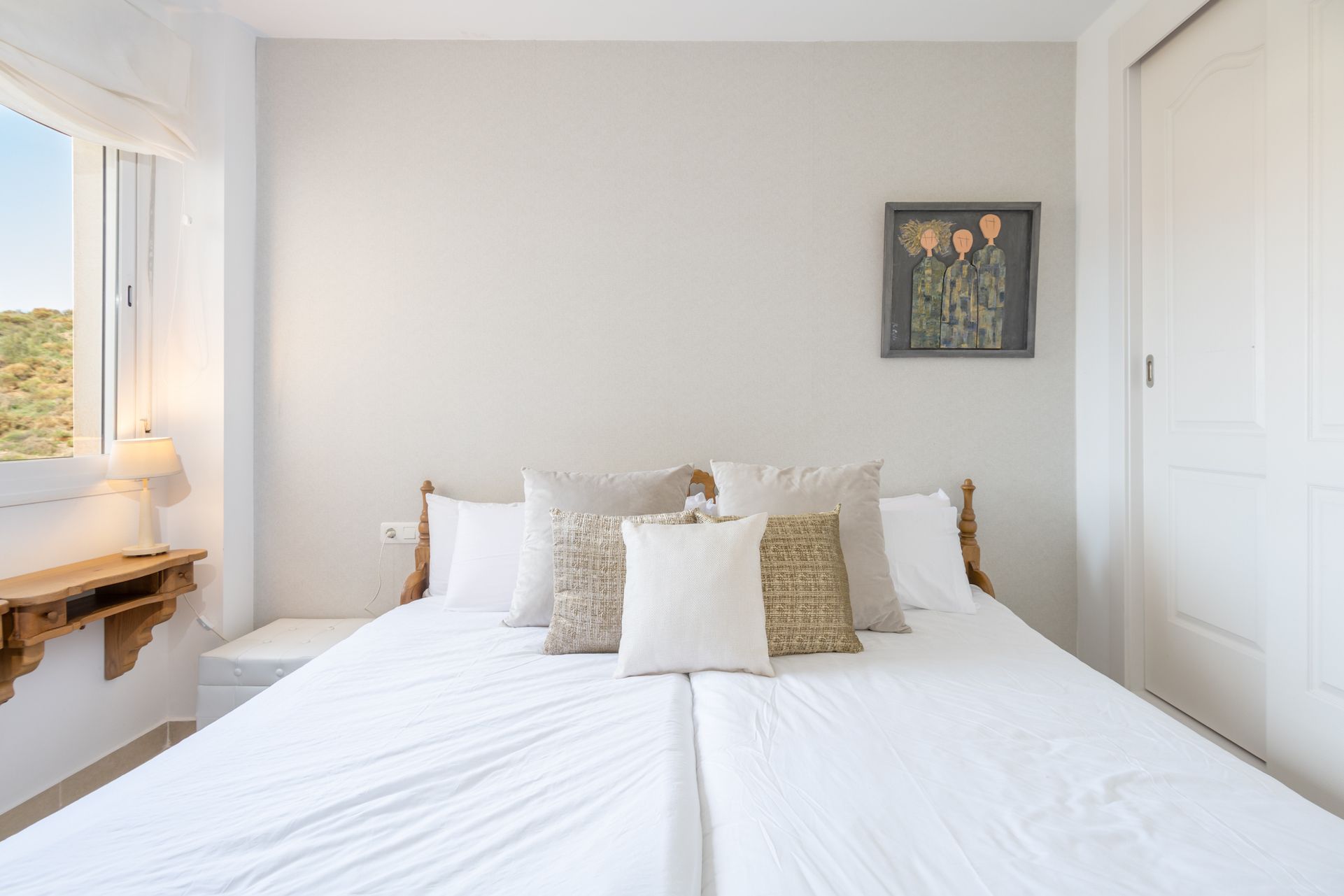 Un dormitorio con cama, almohadas, una mesita de noche, una lámpara y un cuadro en la pared.