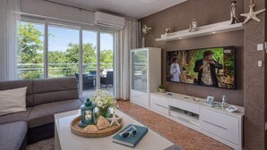 A modern living room with a large flat-screen TV, a sofa, a coffee table with decorative items, and a balcony with a view of greenery. - Sea Breeze Residence Mariella, Umag, Croatia (Umag)
