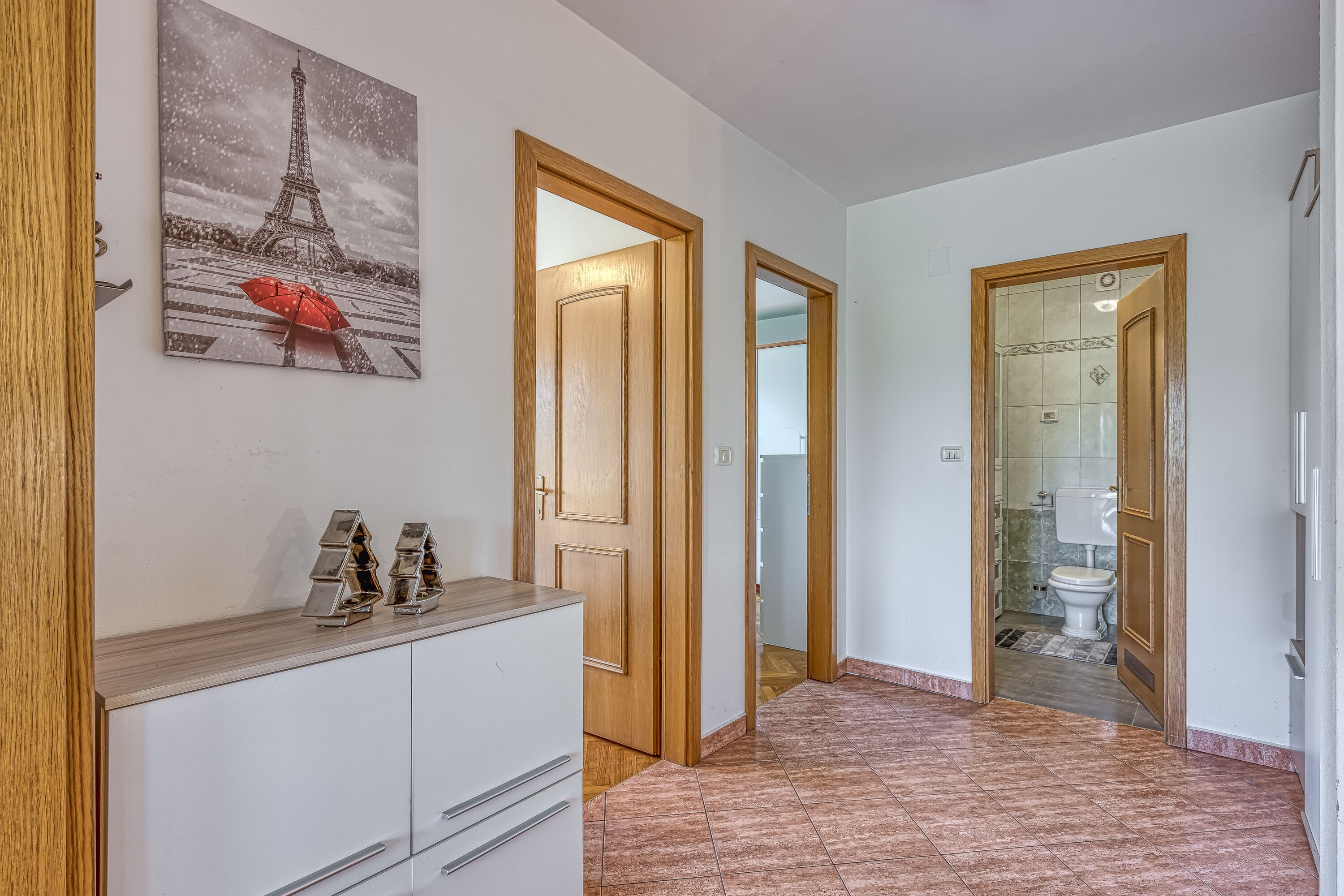A hallway with a wooden cabinet, a painting of the Eiffel Tower, and a bathroom visible through an open door.