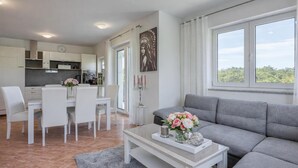A modern living room with a grey sofa, a dining area with white chairs, and a kitchen in the background. - Sea Breeze Residence Mariella, Umag, Croatia (Umag)