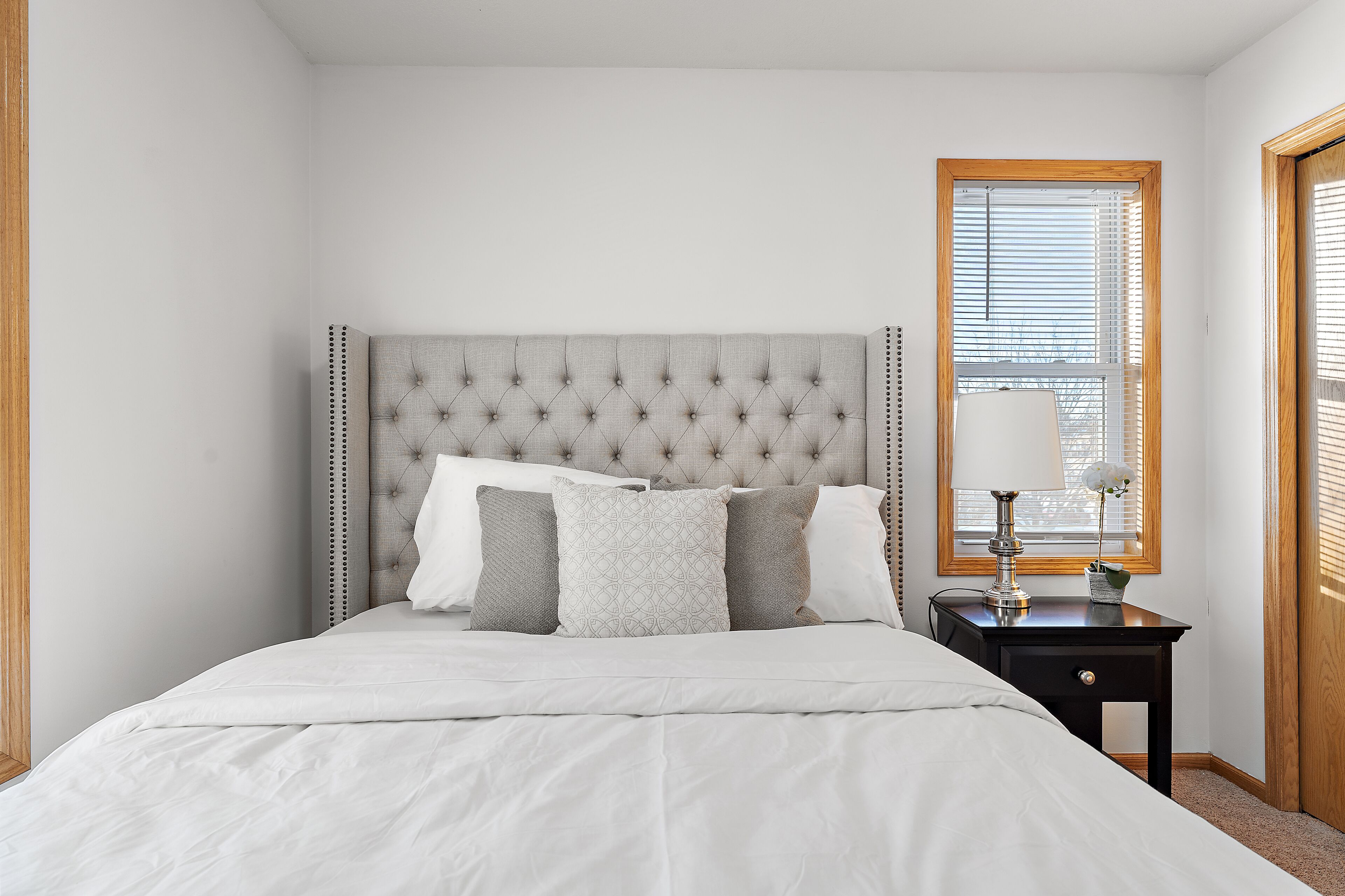Une chambre à coucher avec une tête de lit capitonnée, un lit avec une literie blanche, une table de chevet avec une lampe et une fenêtre avec des stores.