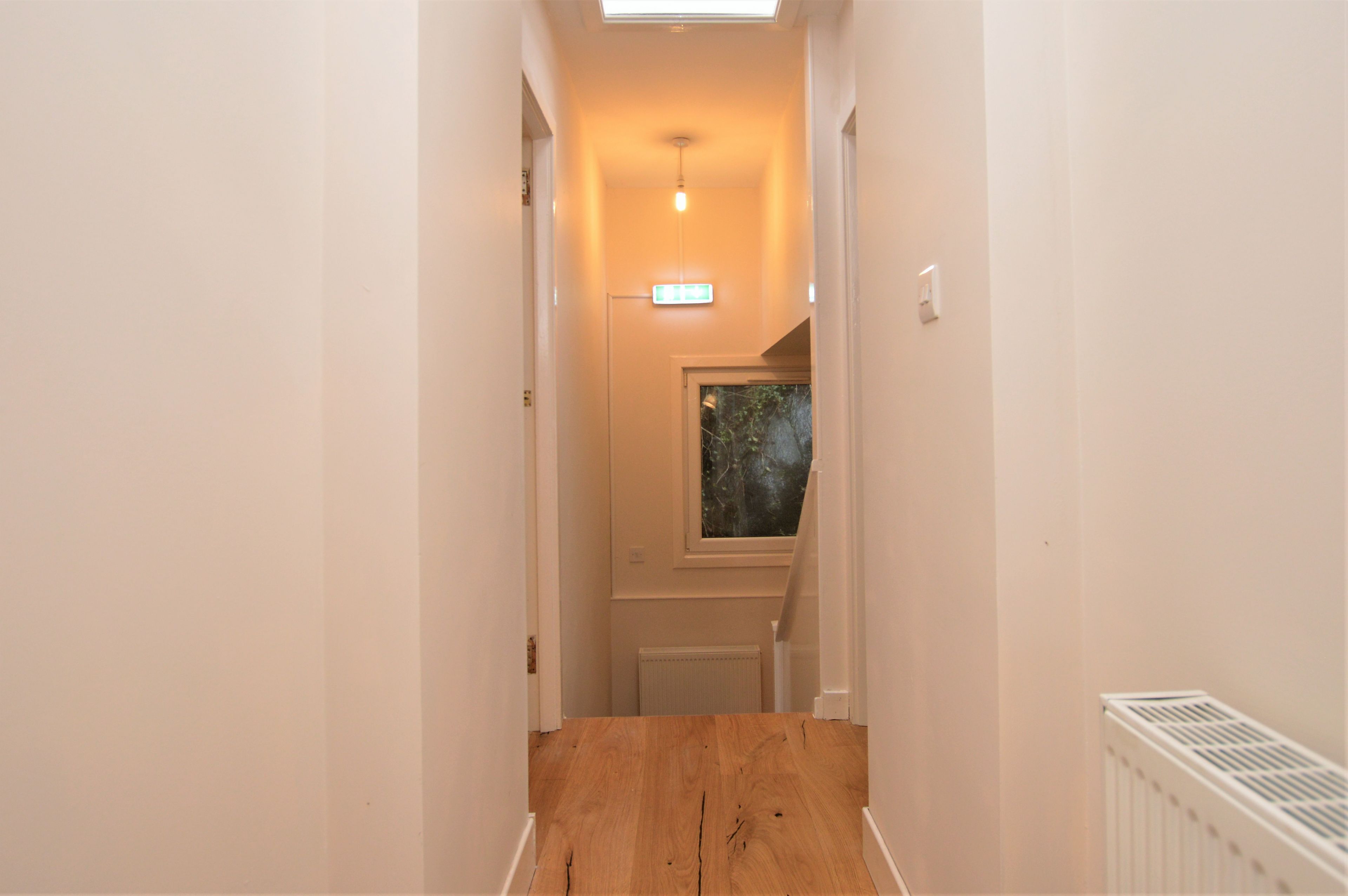 A narrow hallway with a wooden floor, a ceiling light, and a window with a view of trees.