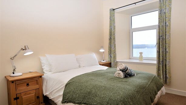 A neatly made bed with a green blanket, two bedside lamps, a wooden nightstand with drawers, and a window with floral curtains. - Pedals and Paddles, waterfront (Dunoon)