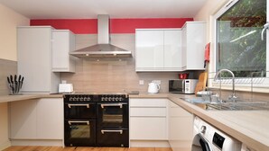 A modern kitchen with white cabinets, a stainless steel range hood, a double oven, a dishwasher, and a window with a view of greenery. - Pedals and Paddles, waterfront (Dunoon)